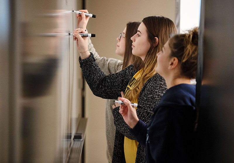 Three students writing on a whiteboard in a UI classroom.