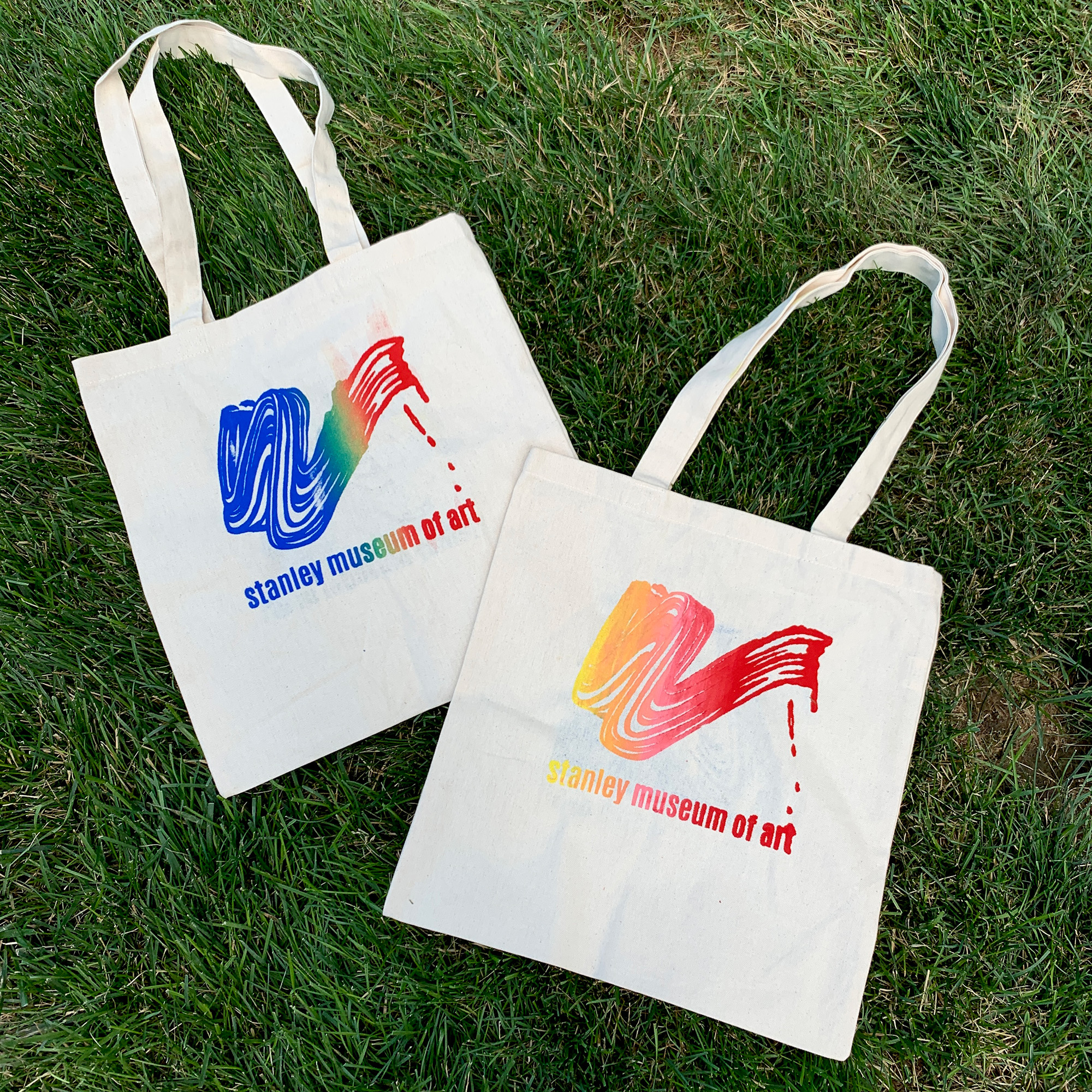 Two hand-printed canvas tote bags lay in the grass. Each has been screenprinted with an abstracted image of a brushstroke and the words "Stanley Museum of Art" in a rainbow of colors.