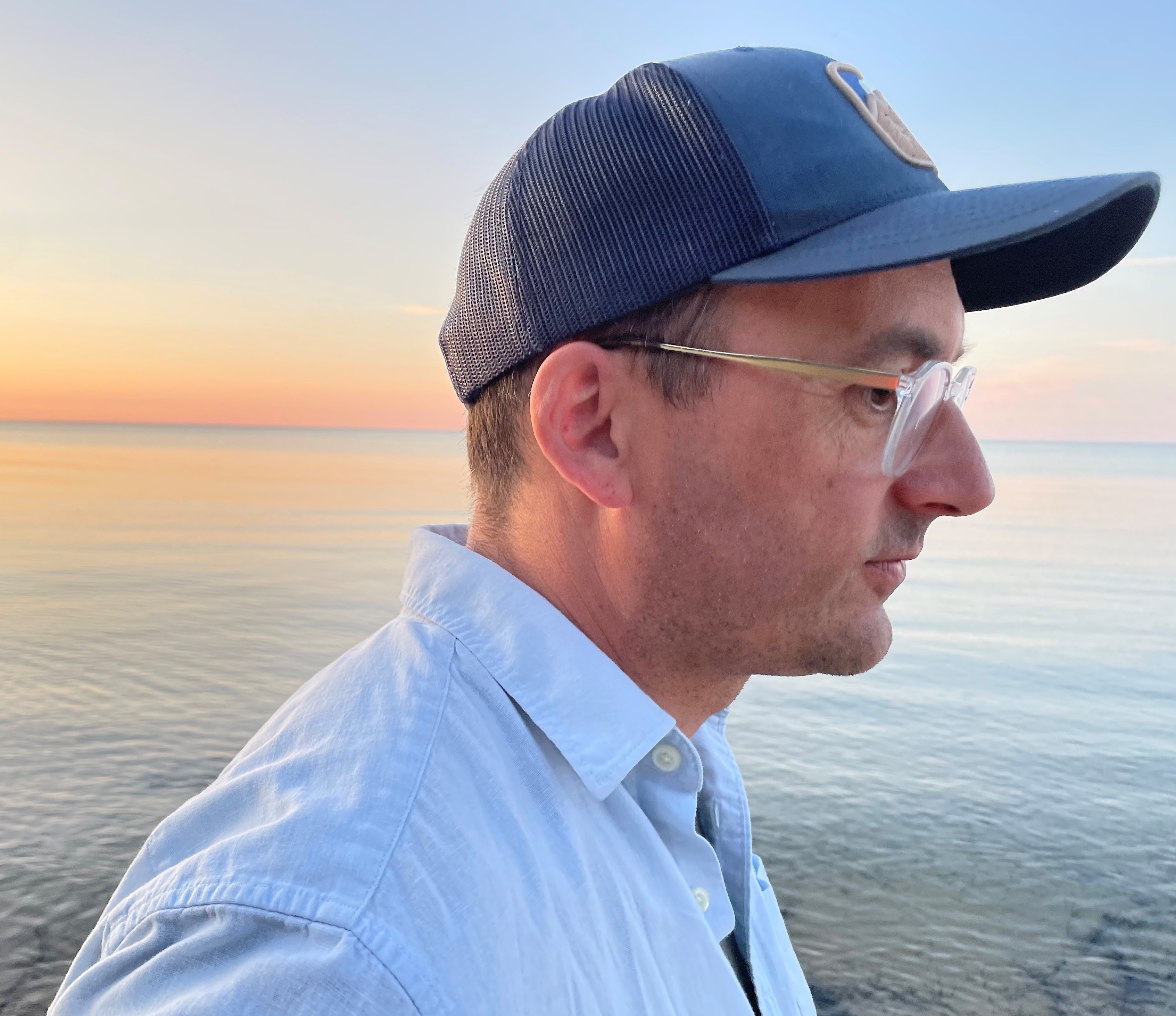 A man wearing a light blue shirt, navy blue ball cap, and glasses is seen in profile. He stands in front of a body of water at sunset.