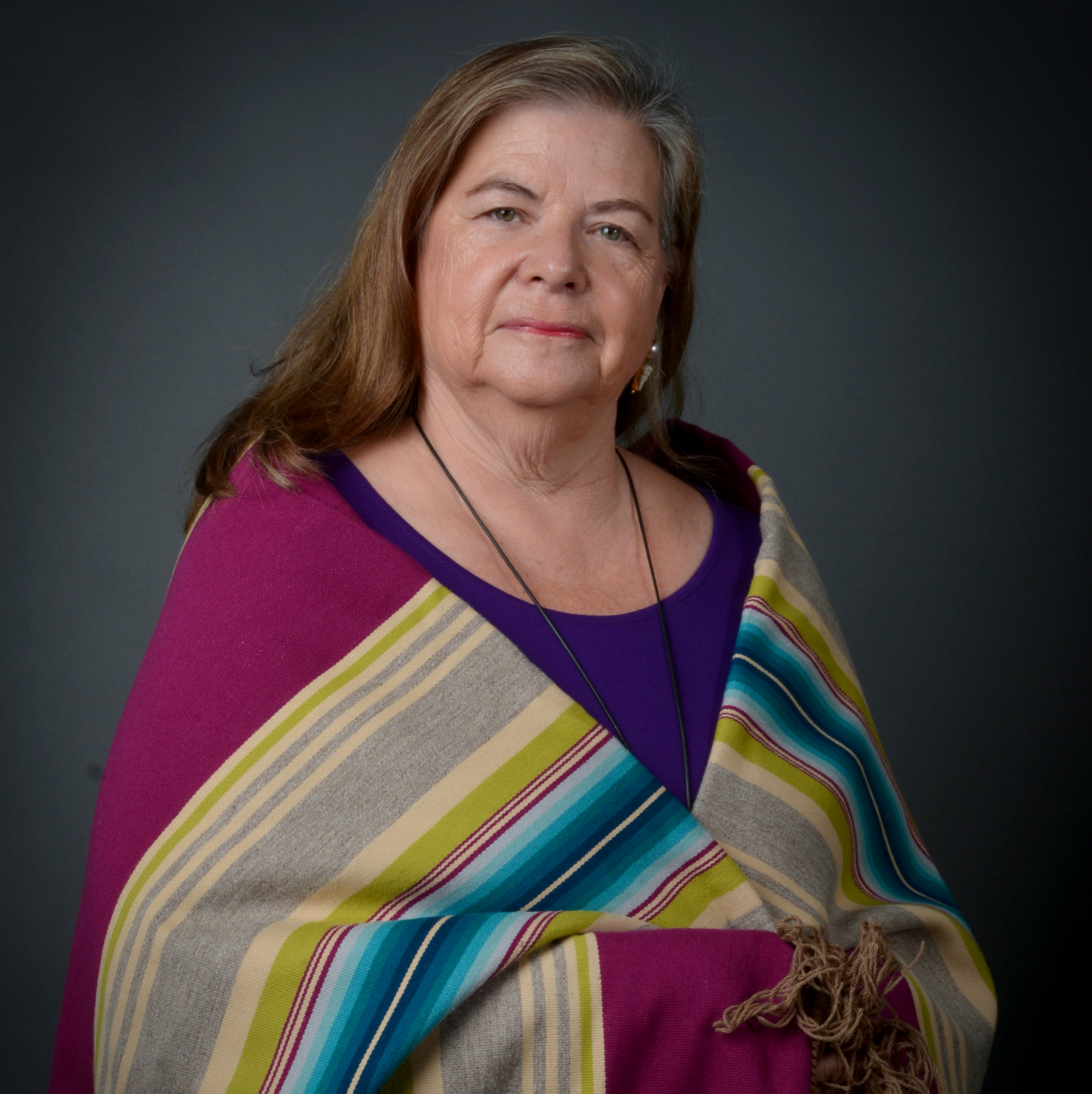 Woman with medium length brown hair wears a purple top and wraps herself in a Native American blanket with multi-colored stripes