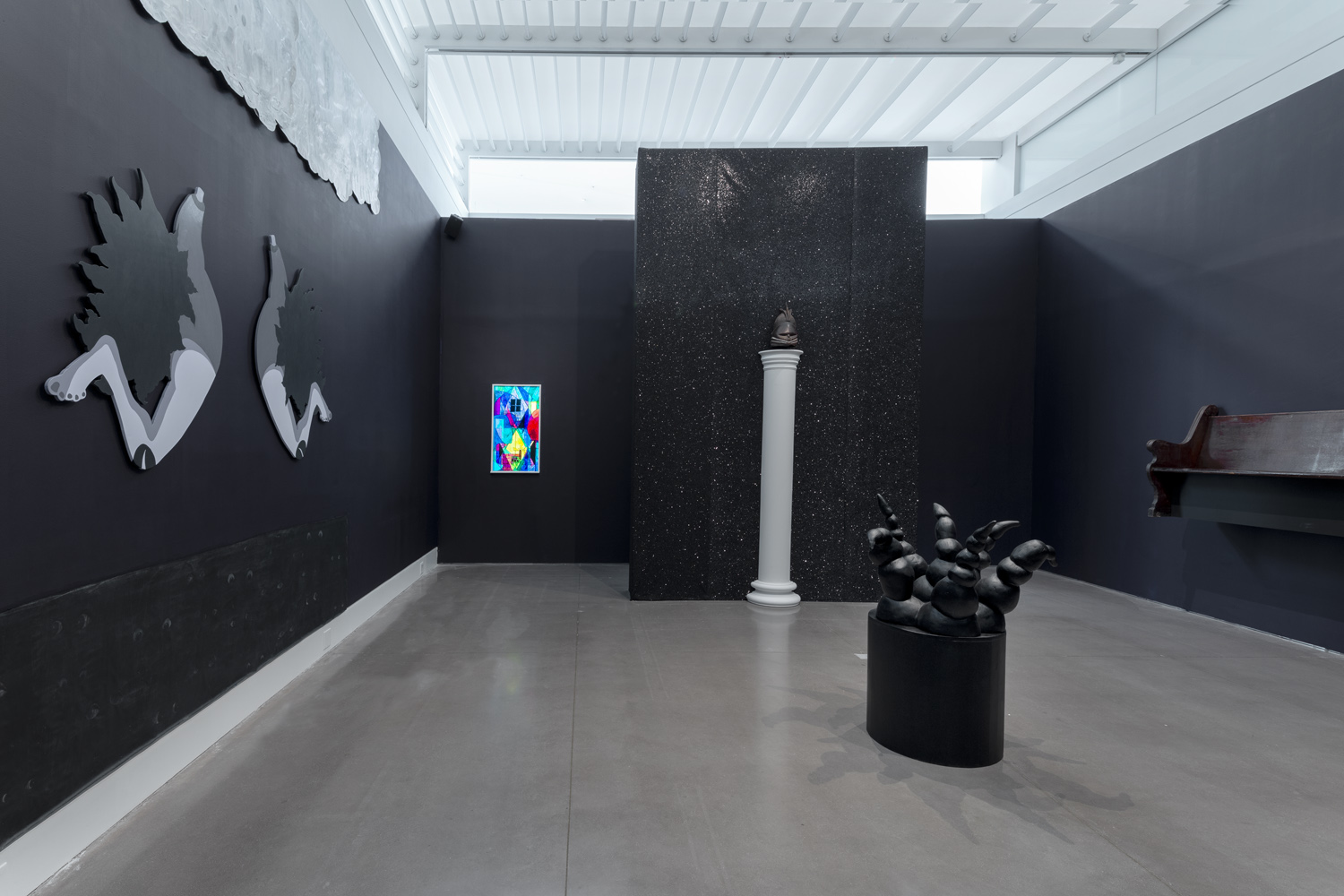 Black walks grey artwork atta ched to wall, colored rectangular artwork on wall, brown wood bench attached to wall, white column with hanging artwork behind it and black art piece on concrete floor 