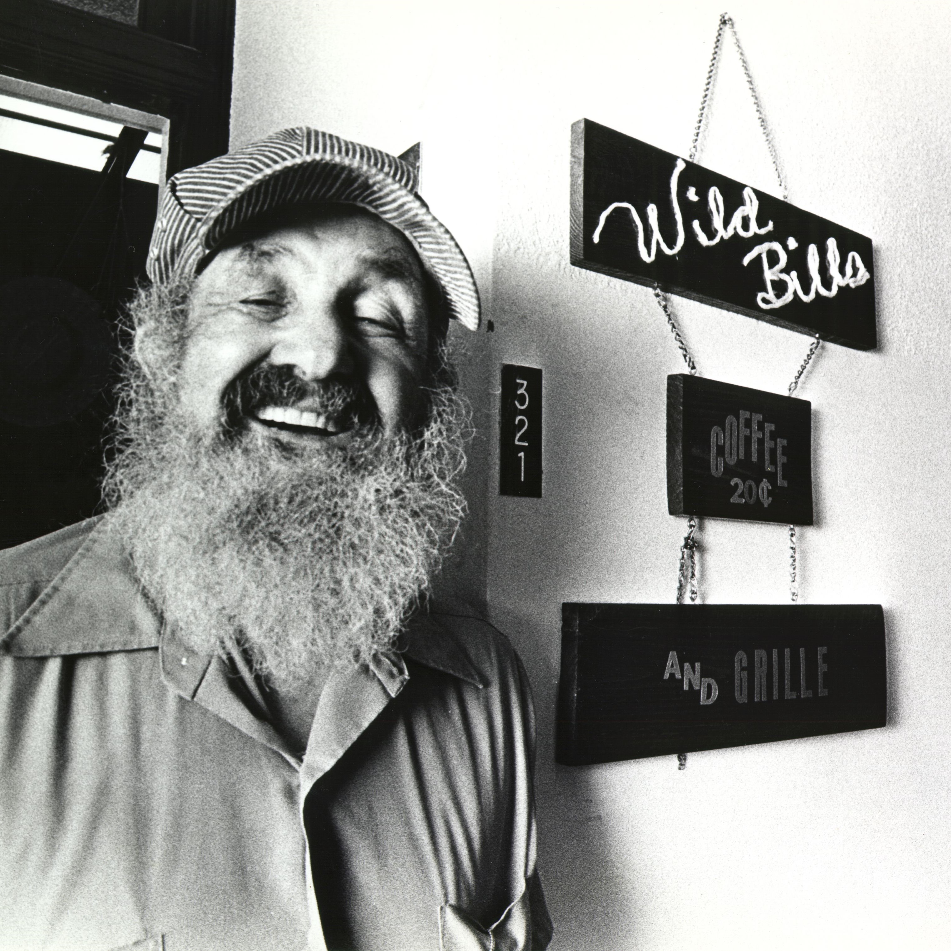 Black and white photo of an elderly bearded man. He is smiling and wears a striped engineer's cap. He stands next to a sign that says Wild Bill's Coffee Shop. The photo is full of joy.
