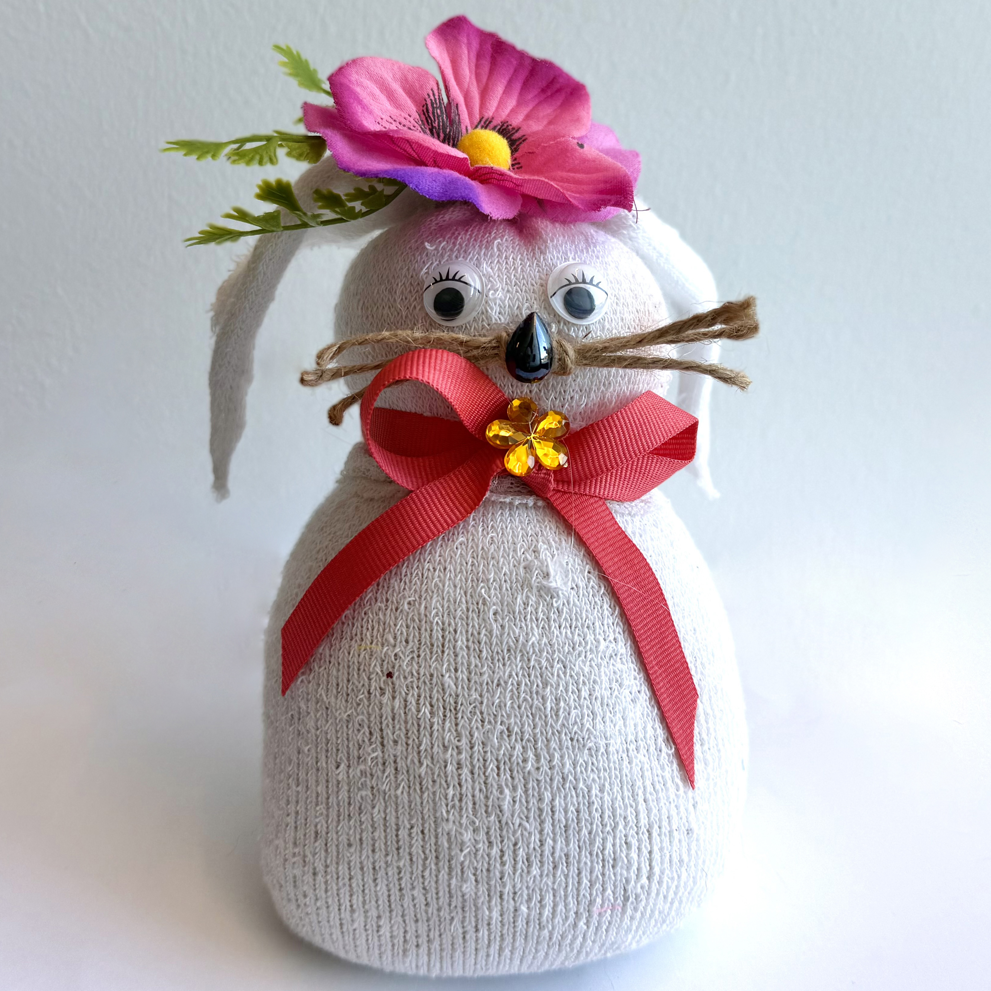 A stuffed sock in the shape of a bunny with a pink flower on top, bead eyes, rope whiskers, and a red ribbon