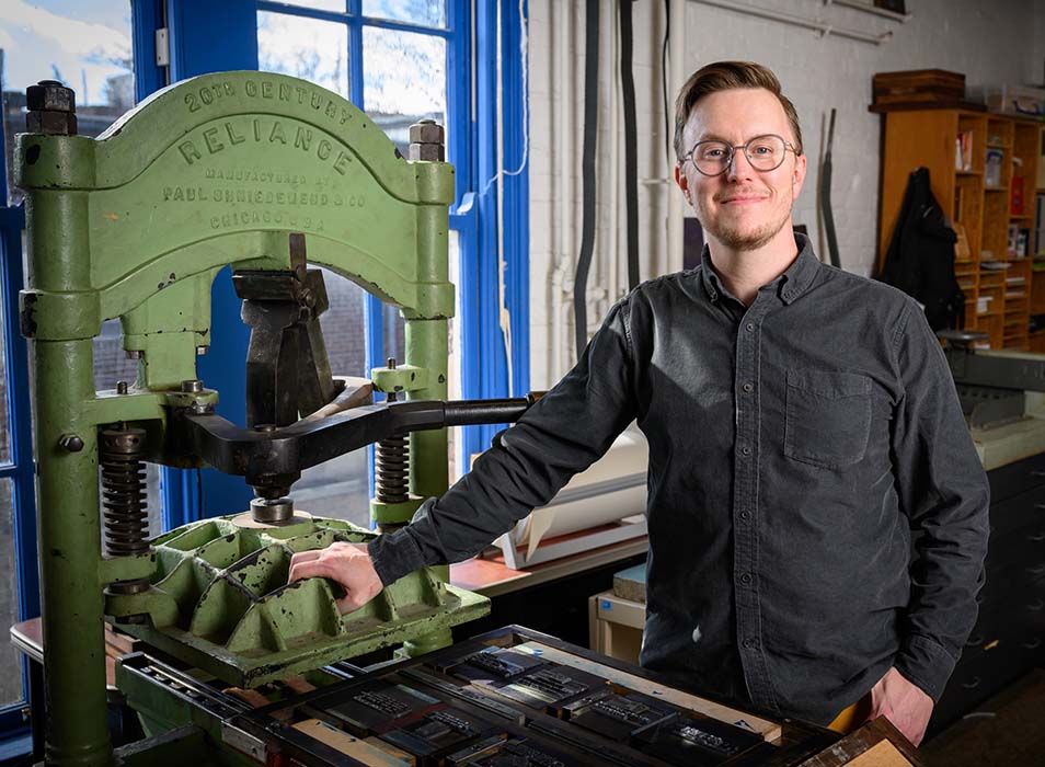 photo of Ryan Cordell wearing a dark gray button down shirt standing in front of a green printing press