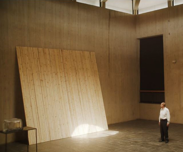 A man stands in a large, cement room looking at a wooden panel that leans against the wall.