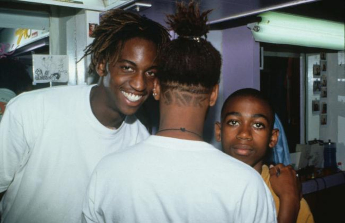 A photo of three young Black men. A man in a white t-shirt smiles at the camera, while another in a white t-shirt looks away from the camera. A much younger boy in an orange t-shirt looks at the camera with a blank expression.