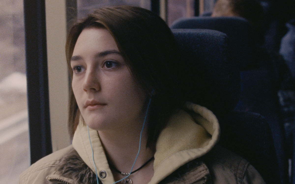 A close-up of a teenage girl in a beige hoodie and brown jacket, listening to music through turquoise earbuds as she looks out the window of a bus.