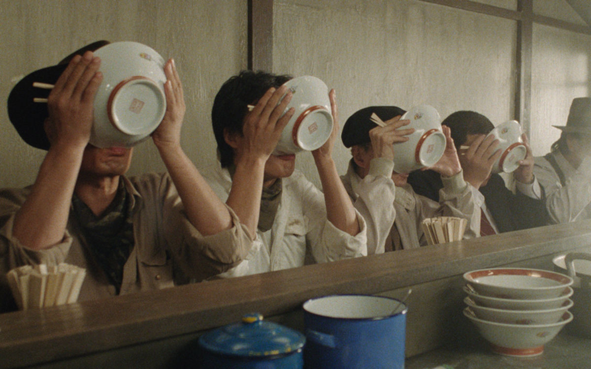 Five men sit in line at a restaurant, all sipping the last broth out of their ramen bowls so that their faces are no longer visible.