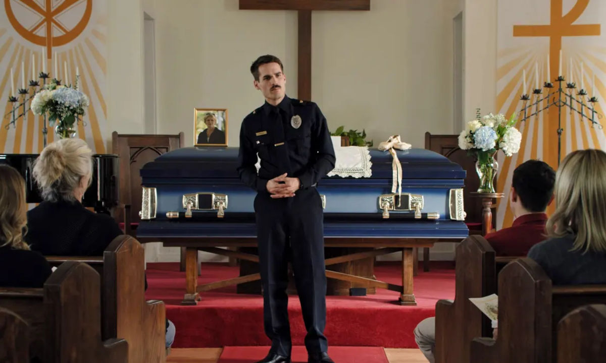 The inside of a church for a funeral. A man in a police uniform stands in front of an audience, a coffin behind him.