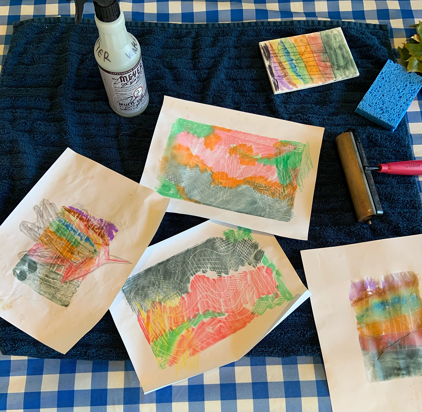 A photo of works of art made by children spread across a table. Various art-making tools and papers lay atop a blue gingham tablecloth. 