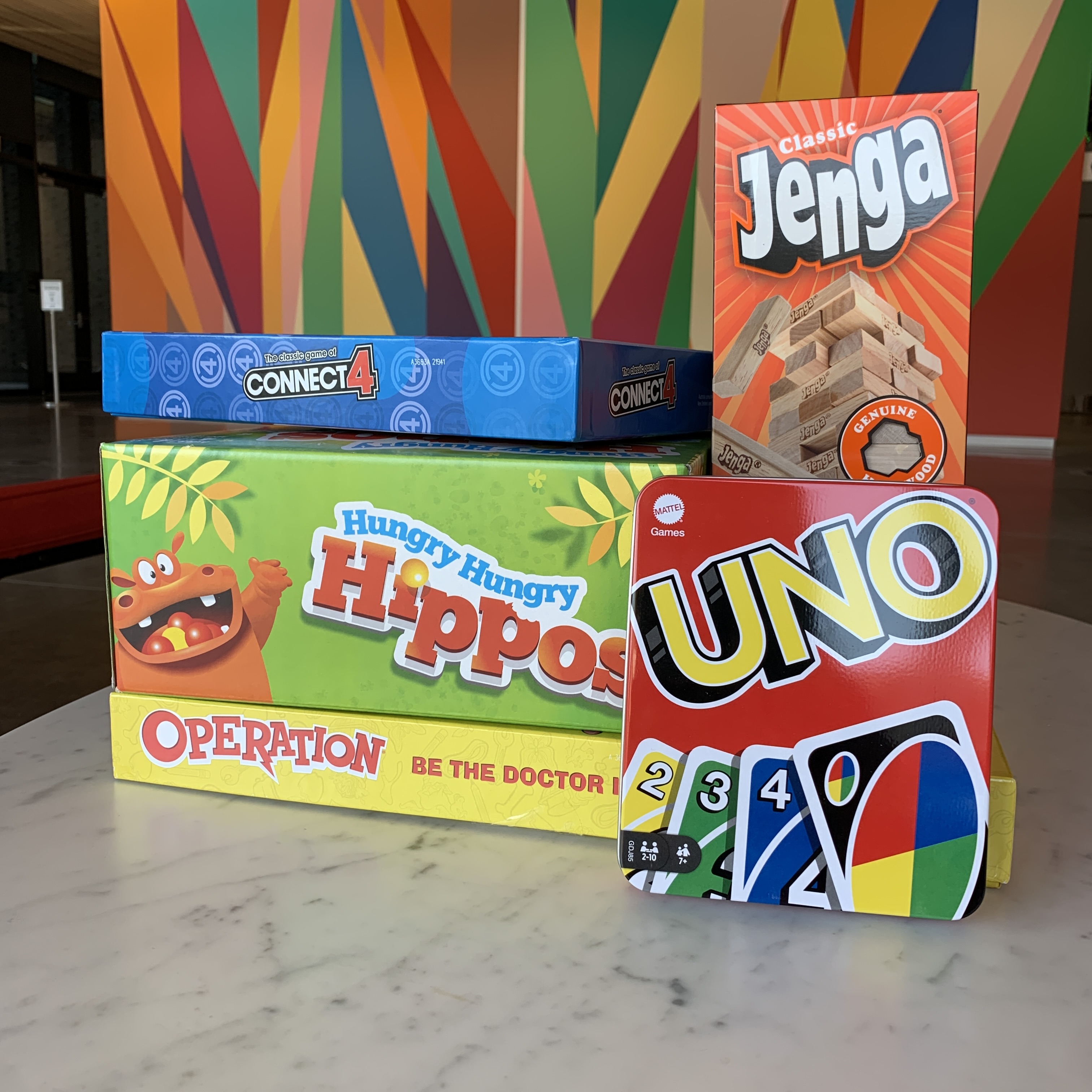 A stack of children's board games rest atop a marble table: Jenga, Hungry Hippos, Operation, Connect 4, and Uno. In the background, you can see the rainbow colored mural in the Stanley Museum of art Lobby.