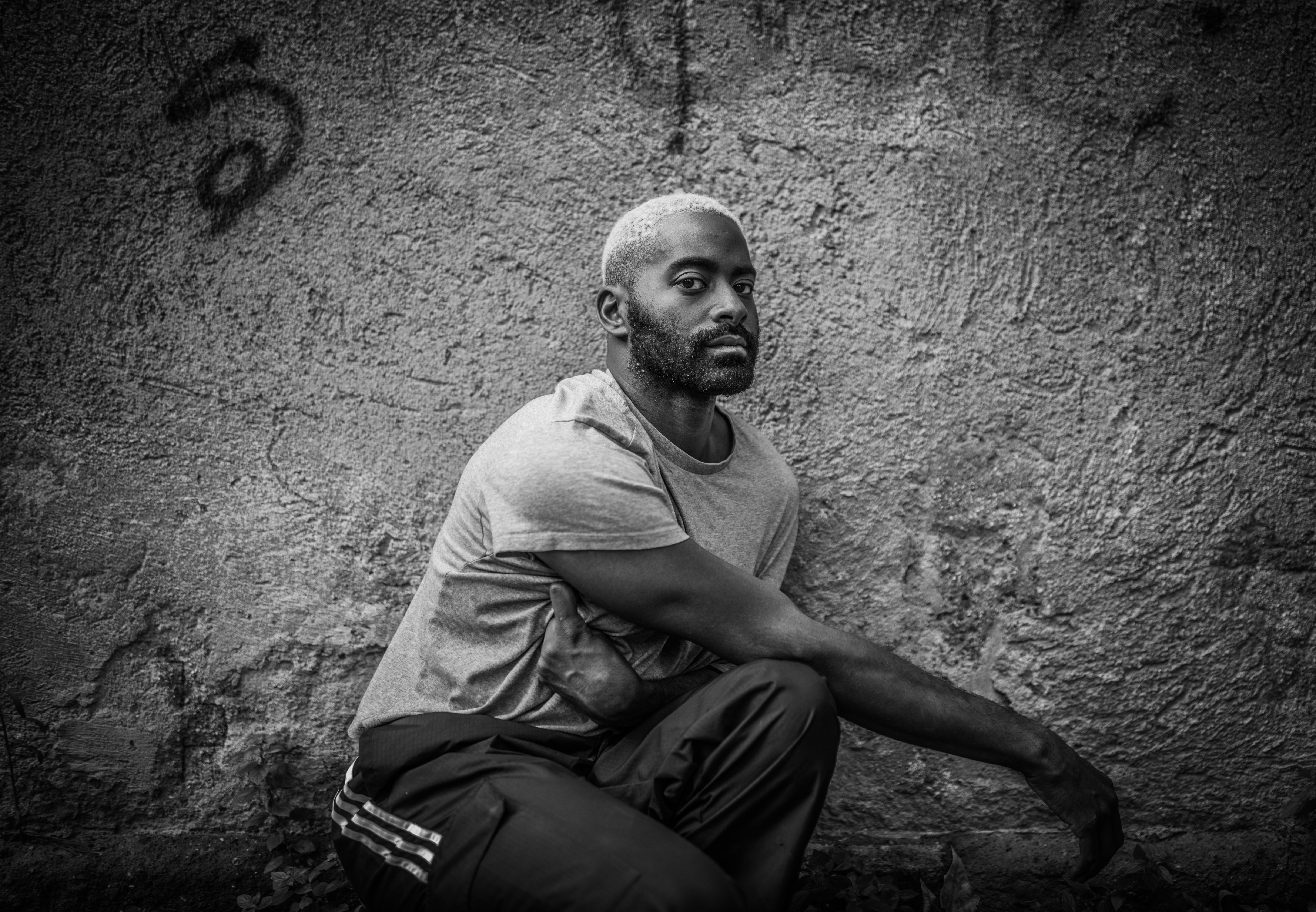 Black and white photo of Jerron, dark skinned, wearing a light grey t-shirt with bleached hair and a dark beard. Against a textured wall with faint graffiti.  