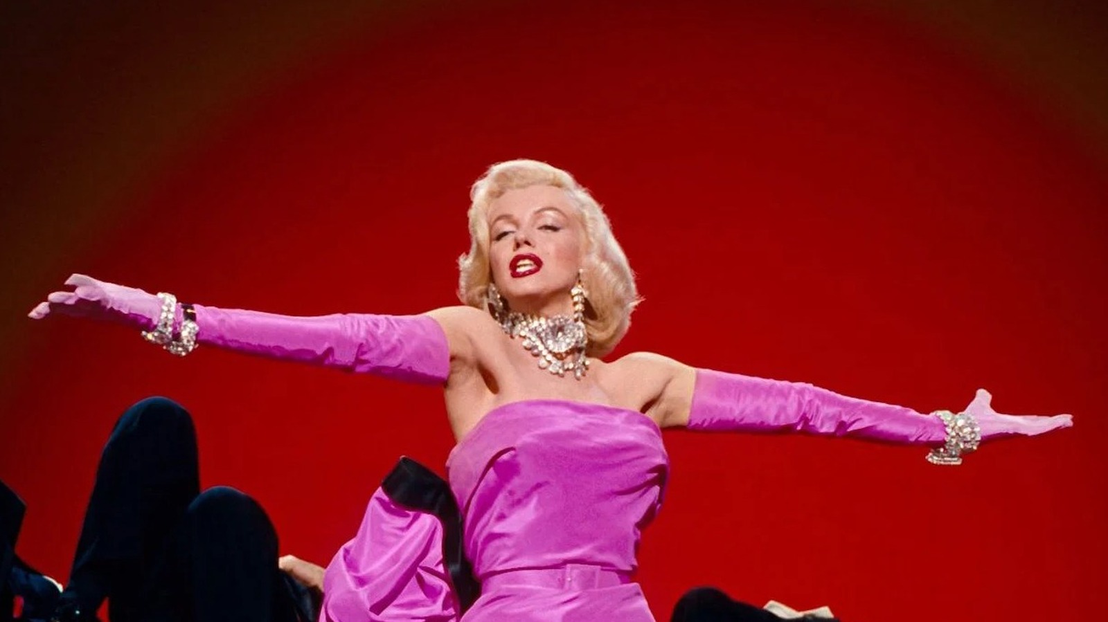 A blonde woman in a bright pink dress and bright pink gloves, with thick diamond jewelry along her neck and wrists, sings with arms outstretched in front of a red background.