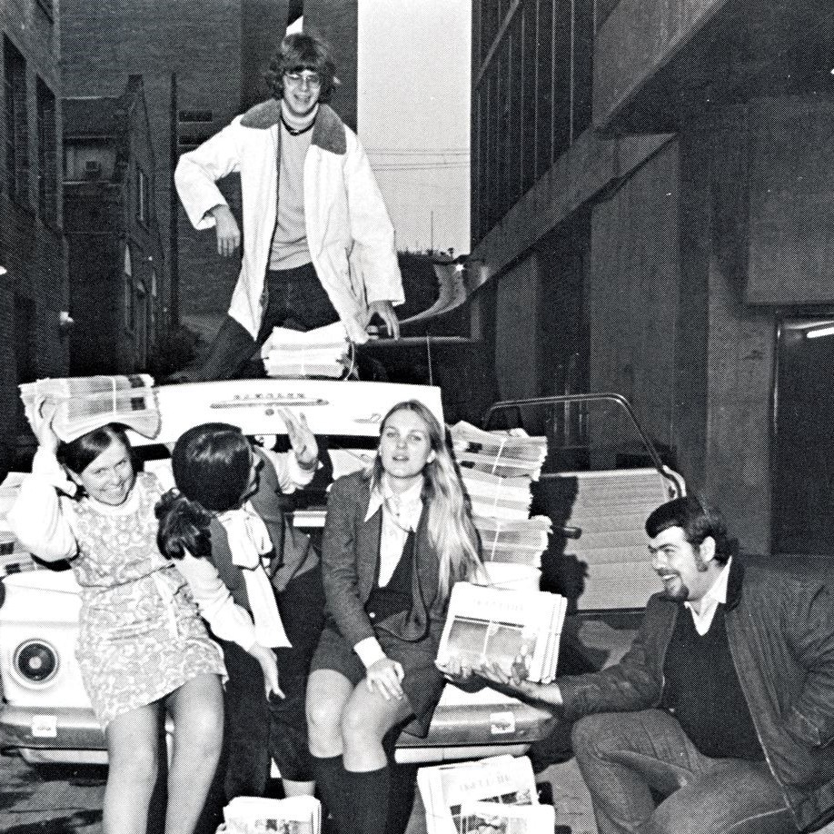 Five students sit on or near the trunk of a car in an alleyway in 1971. This candid photo shows them laughing and talking while holding bundles of student-produced newspaper The Light Eater.