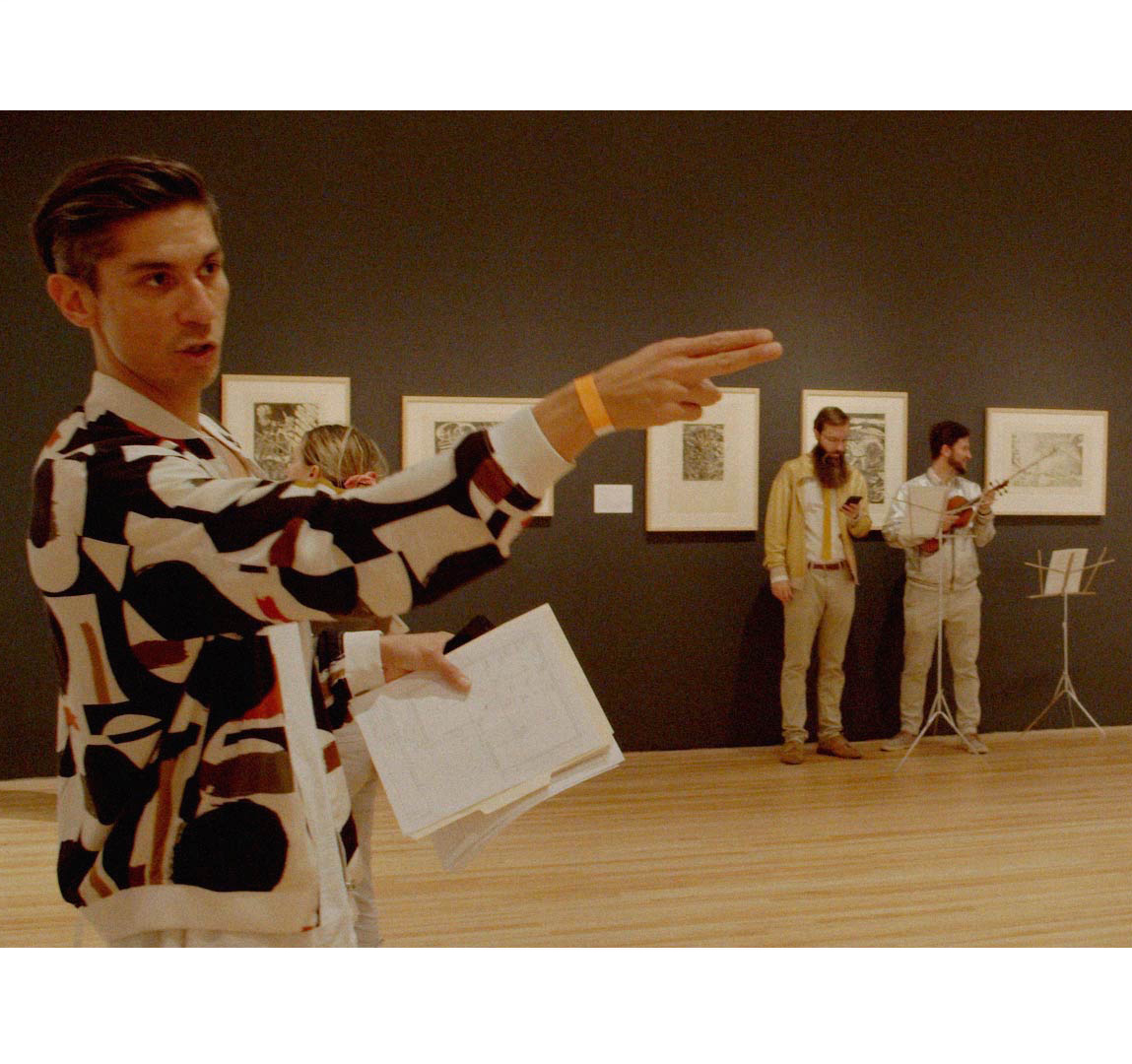 A man with short, dark hair wearing a bomber jacket with bold, geometric patterns, gestures with his arm. He stands in an art gallery with framed artworks on the wall behind him.