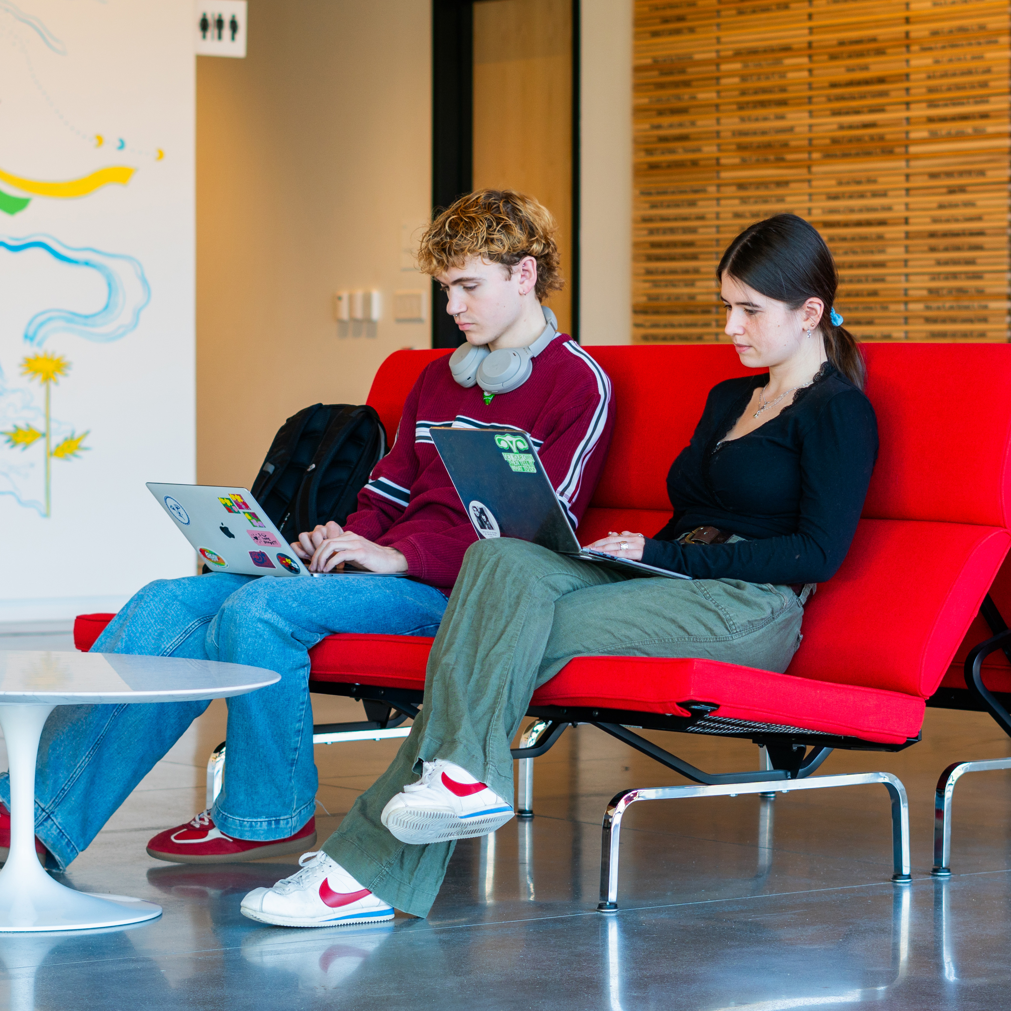 Two people seated on a red couch working on laptops