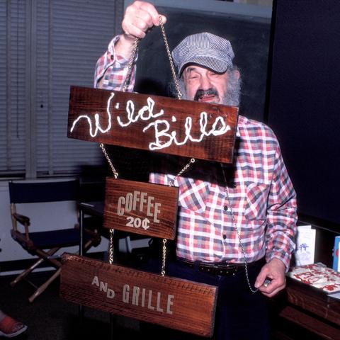 Bill Sackter, an elderly man with a big bushy gray beard, smiles as he holds up a wooden sign that says Wild Bill's Coffee 25 cents and grill. He is wearing a plaid shirt and a train engineer-style cap.