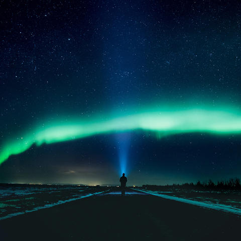 a person standing in a road looking at green northern lights in a starry sky