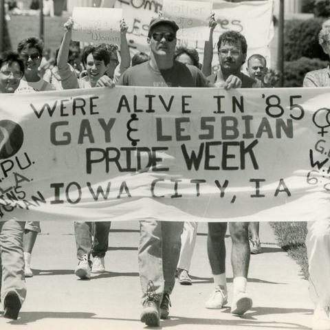 Several smiling people walk behind a banner that says "We're alive in 85. Gay and lesbian Pride Week, Iowa City, Iowa.