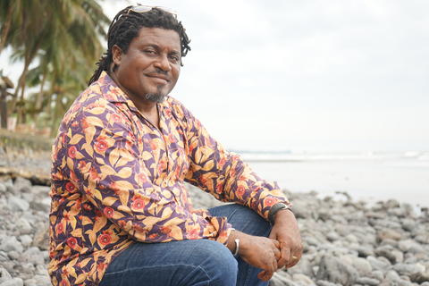 A Black man wearing a shirt with bright floral print sits on a beach
