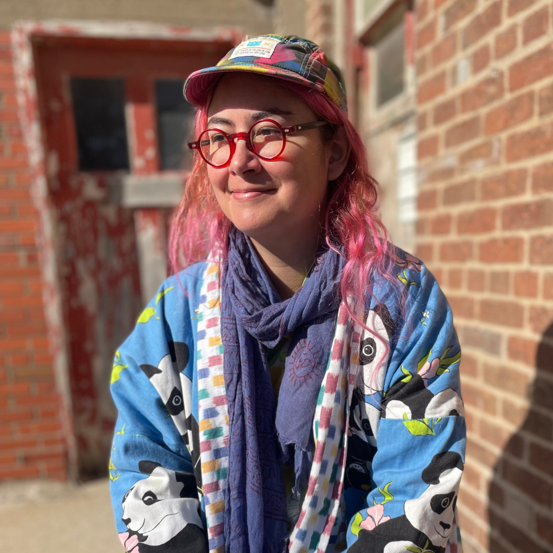 Headshot of artist Lilah Ward. Lilah stands outdoors, in front of a brick wall. She is smiling and she stares off to the left, and is wearing red circular-framed glasses and her hair is partially pink. 