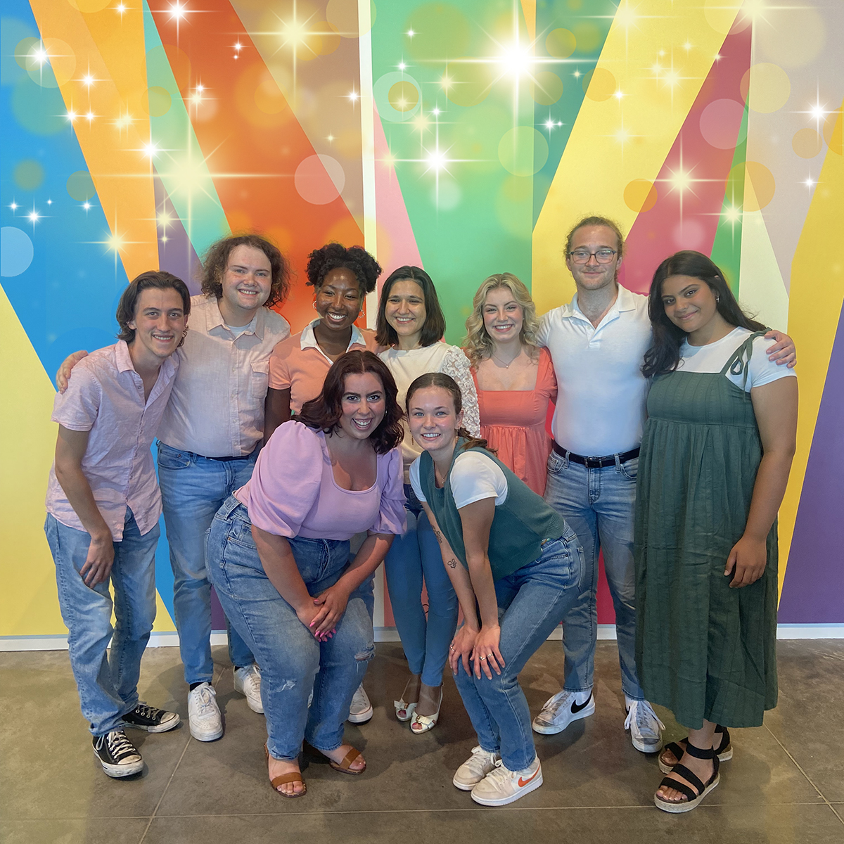 Group of people standing in front of a brightly colored wall. There are golden sparkles superimposed over their heads