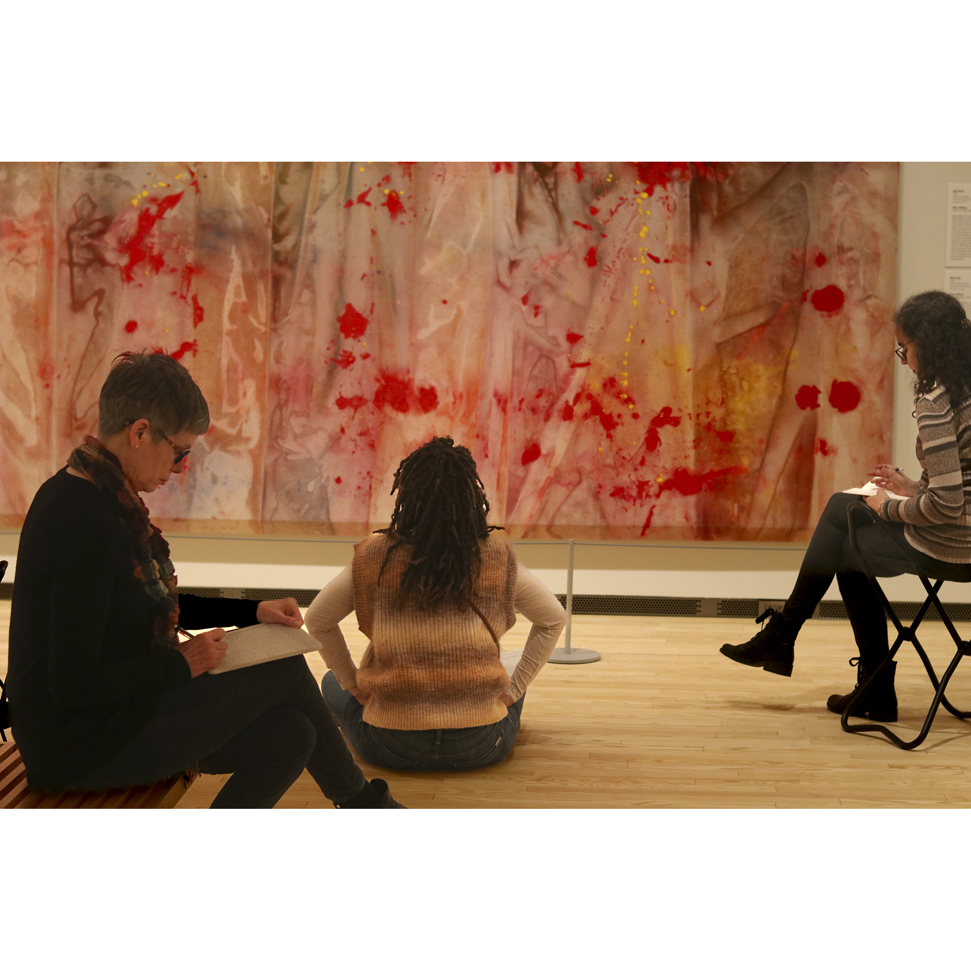Three women are seated and writing in notebooks in a museum gallery